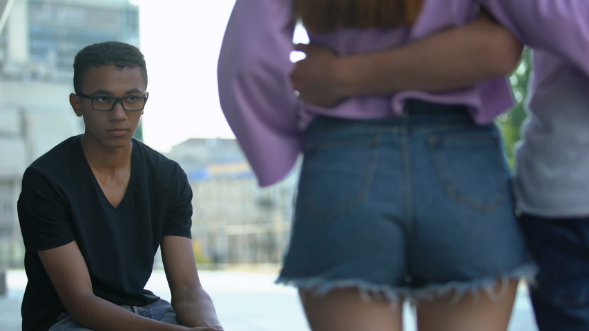 Insecure african teenager in eyeglasses feeling lonely looking at hugging couple