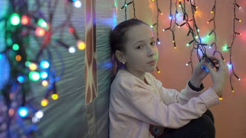 Portrait of a sad little girl on the background of flashing colorful Christmas lights. She sits among the small, colorful garlands, and looking at the camera. Shallow depth of field. Close-up. 4K