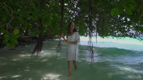 Beauty young girl in white dress walking in foaming sea, surrounded by lianas, tropical trees, plants. Maldives. Bounty paradise. Super slow motion. Freedom mood.