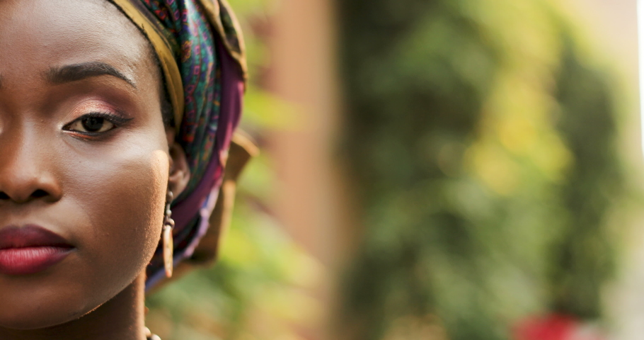 Close up of the half face of the young African beautiful woman with a traditional scarf on the head looking straight to the camera and smiling. Portrait.