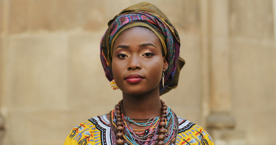 Close up of the young African attractive woman in the traditional clothes and with scarf over her head looking straight to the camera and smiling on the wall background. Portrait.