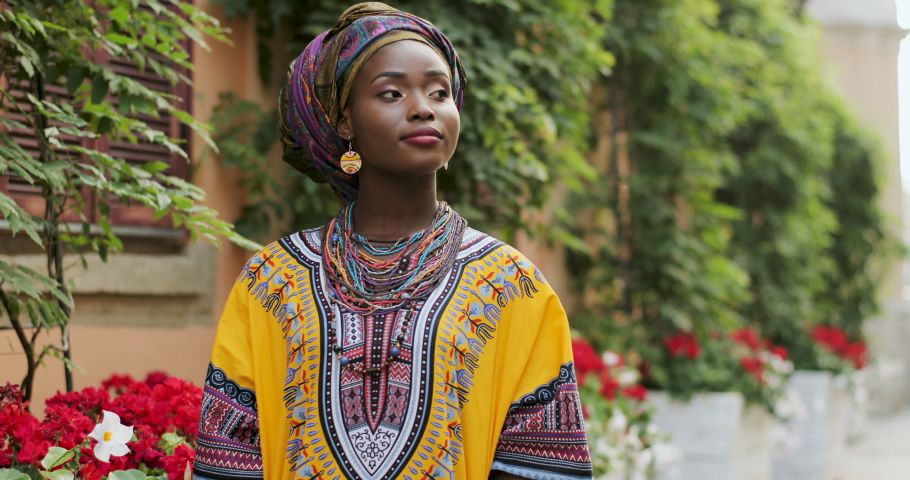 Portrait shot of the beautiful and happy Afrcan woman in the traditional outfit standing in the cozy courtyard with flowers and smiling to the camera. Outdoor.