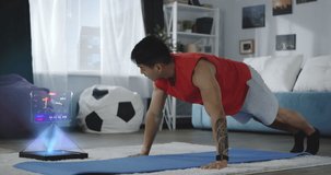 Young asian athlete doing push-ups at home using holographic futuristic computer with holographic screen displaying exercise in app