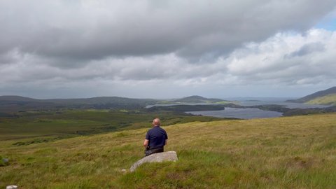 Old man sitting on boulder overlooking Irish spectacular countryside sights of into distance.