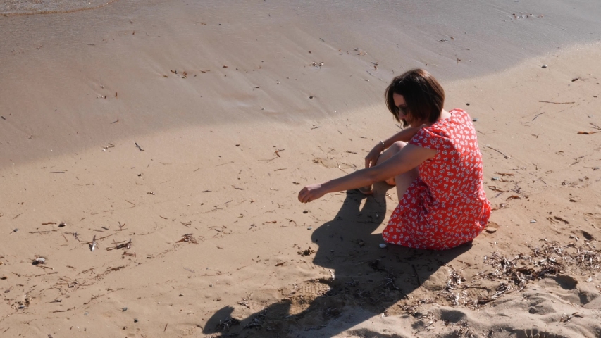 Young attractive girl in red dress sitting on yellow sand near sea. 
