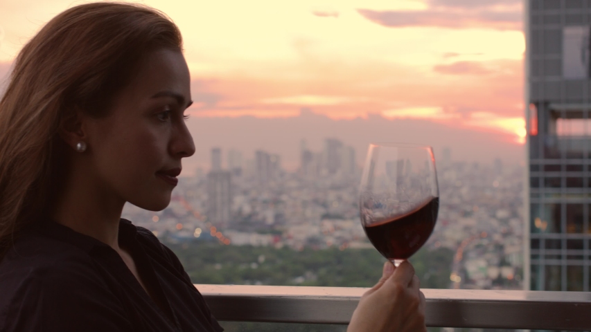 young woman tastes and drinks red wine with in the background a sunset over the city, Manila Philippines