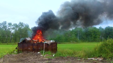 Thick dark smoke billowing from a burning dumpster full of garbage on rural farmland.