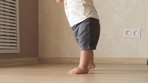 Closeup of baby tiny barefoot legs doing first steps and walking on floor at home without any help. Slow motion