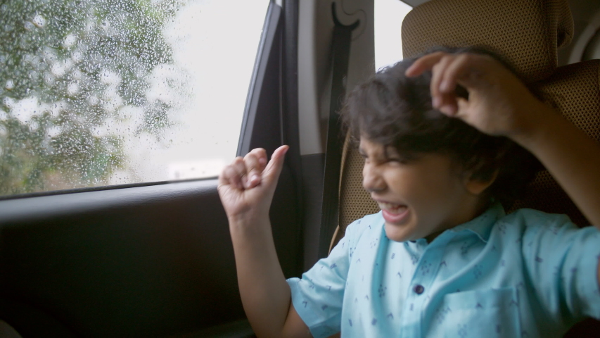 Happy Indian child enjoying music while traveling back home in a car - childhood concept. A handsome young kid listening and dancing to his favorite songs, sitting at the backseat of a car in casua...