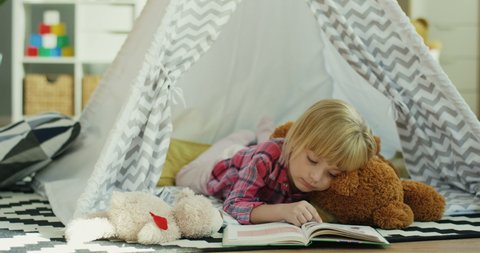Close up of the blond cute and pretty Caucasian little girl hugging a toy teddy bear and reading an interesting book while lying in the wigwam in her nice rom.