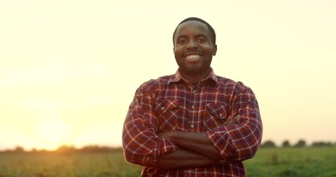 Portrait of the handsome young African American man farmer standing in the field on the sunset, smiling to the camera and crossing his hands in front of him.