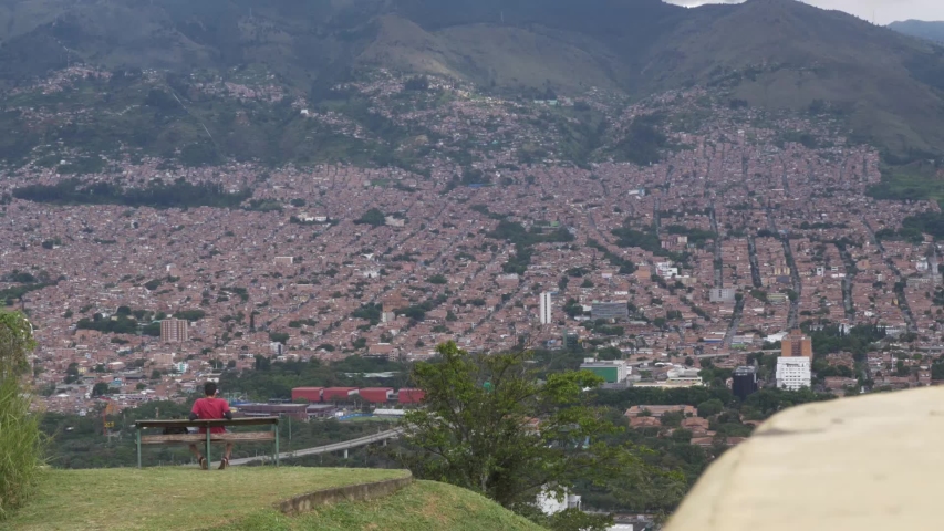 Wide shot of man in red shirt sitting on bench looking out into Medellin and its surrounding mountains.