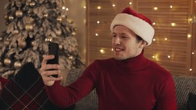 Cheerful Attractive Caucasian Man Wearing Santas Hat Siiting on the Sofa near Christmas Tree Using his Smartphone to Make a Video Call or Video Message Concept of Holidays and New Year Side View.