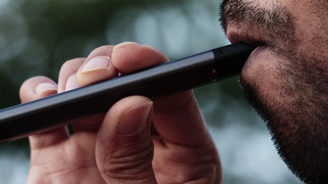 Young male with beard puffs on a vape pen device few drags outdoors, extreme close up