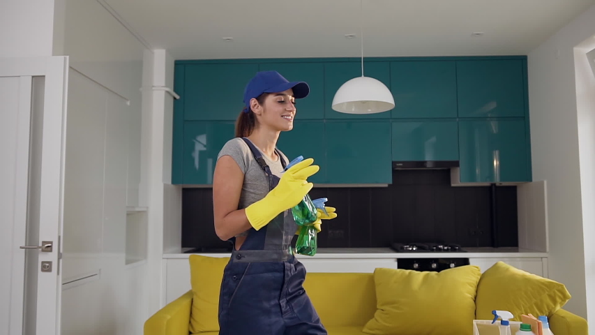 Cheerful young woman from cleaning service dressed in workwear and rubber gloves keeping in hands cleaning agents and spraying them under her dancing