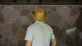 A man in a shirt and a straw hat is dancing against a stone wall. On the face is a beard. Average age. The farm. Country life. Portrait to the waist. 4K video