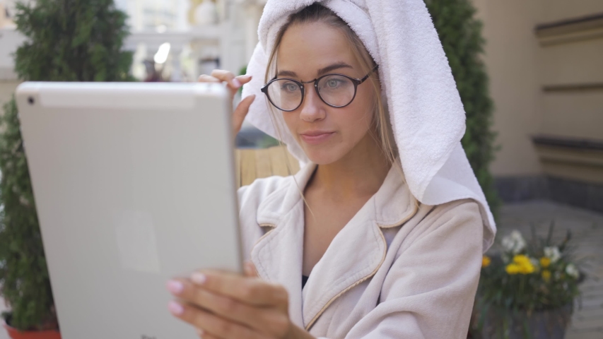 Portrait of fun cute young woman in glasses and bathrobe with towel on head looking at tablet sitting on backyard. Confident girl enjoying sunny day outdoors. | Shutterstock HD Video #1036941431