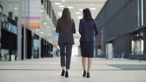 Rear view full length follow shot of two businesswomen in formal suits and high heels walking down office building lobby together and talking. Middle aged woman carrying laptop computer
