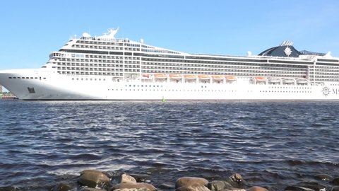 RIGA/LATVIA - MAY 18 2019: Huge white cruise tourist liner sails on crystal deep blue water into harbor past small speed boat close view on May 18 in Riga