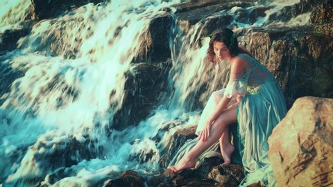 A young attractive woman in a wet water color dress sits on stones and strokes her bare leg. Tender, fragile, slim, river nymph, resting near a tropical waterfall. Bright sunny day. Dreamy Princess.