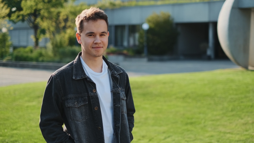 Portrait of the cute Caucasian teenage boy smiling to the camera, then his pretty girlfriend coming, embracing him and they posing together outside at some building of college.