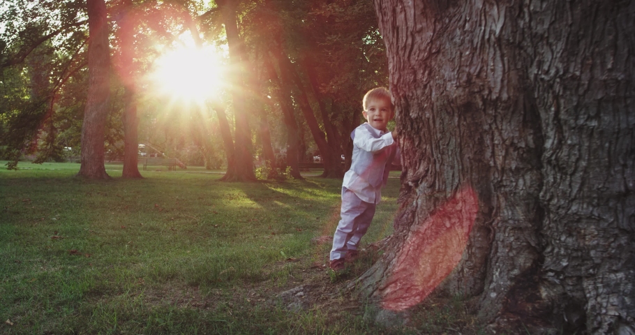 Very cute and funny boy hiding out beside of a big tree in the middle of the park he wearing a beautiful stylish suit