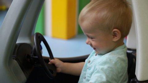 Young driver. Child closes the door in a toy car and controls the steering wheel. Baby boy, one year old. Side view. Children's play center.