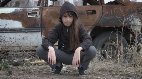 Gangster girl. Child in the hood sitts on her haunches in loneliness near a burnt car.