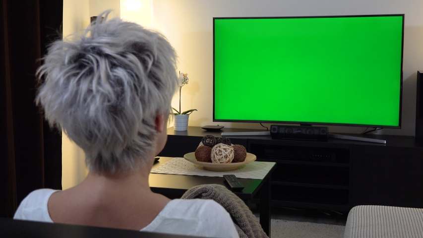 A middle-aged woman sits on a sofa in an apartment living room and watches TV with a green screen - view from behind