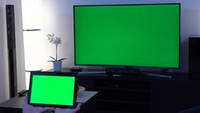 A woman holds out a tablet with green screen in front of camera in a living room with a TV with green screen