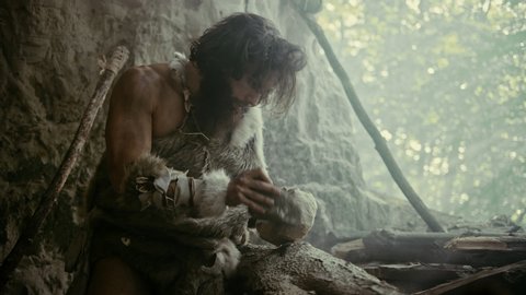 Primeval Caveman Wearing Animal Skin Hits Rock with Sharp Stone and Makes Primitive Tool for Hunting Animal Prey. Neanderthal Using Hand axe to Create first Millstone. Slow Motion Shot