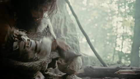 Primeval Caveman Wearing Animal Skin Hits Rock with Sharp Stone and Makes Primitive Tool for Hunting Animal Prey. Neanderthal Using Hand axe to Create first Wheel. Slow Motion Shot