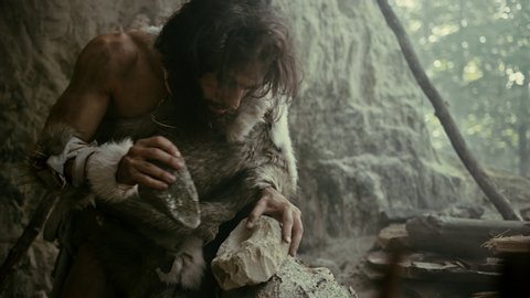 Primeval Caveman Wearing Animal Skin Hits Rock with Sharp Stone and Makes First Primitive Tool for Hunting Animal Prey. Neanderthal Using Flint Rock. Dawn of Human Civilization. Slow Motion Closeup 