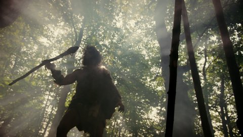 Primeval Caveman Wearing Animal Skin Holds Stone Tipped Spear Looks Around Prehistoric Forest, Ready to Hunt Animal Prey. Neanderthal Going Hunting into the Jungle. Low Angle Slow Motion Arc Shot