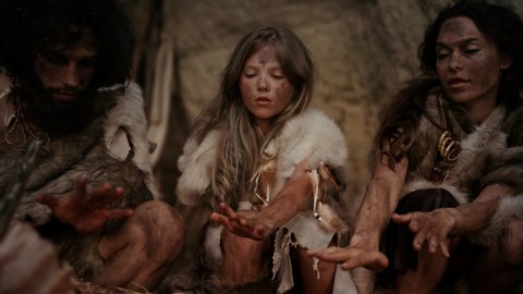 Tribe of Prehistoric Hunter-Gatherers Wearing Animal Skins Live in a Cave at Night. Neanderthal or Homo Sapiens Family Trying to Get Warm at the Bonfire, Holding Hands over Fire, Cooking Food