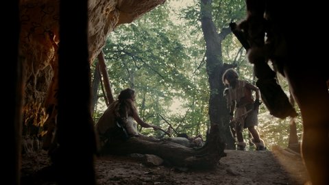 Tribe of Hunter-Gatherers Wearing Animal Skin Live in a Cave. Men Brings Animal Prey from Hunting, Female Cooks Food on Bonfire, Girl Drawing on Wall. Neanderthal or Homo Sapiens 