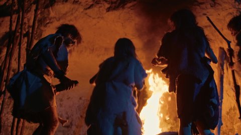 Tribe of Prehistoric Hunter-Gatherers Wearing Animal Skins Dance Around Bonfire Outside of Cave at Night. Neanderthal / Homo Sapiens Family Doing Pagan Religion Dancing Near Fire Back View Slow Motion