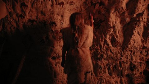 Primitive Prehistoric Neanderthal Child Wearing Animal Skin Draws Animals and Abstracts on the Walls at Night. Creating First Cave Art with Petroglyphs, Rock Paintings. Back View Following Shot