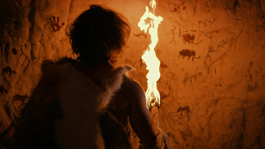 Primeval Caveman Wearing Animal Skin Standing in His Cave At Night, Holding Torch with Fire Looking at Drawings on the Walls at Night. Cave Art with Petroglyphs, Rock Paintings. Back View Royalty-Free Stock Footage #1037019383