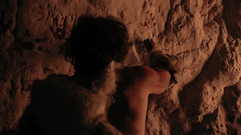 Primitive Prehistoric Neanderthal Wearing Animal Skin Draws Animals and Abstracts on the Walls at Night. Creating First Cave Art with Petroglyphs, Rock Paintings Illuminated by Fire.Back View Zoom Out