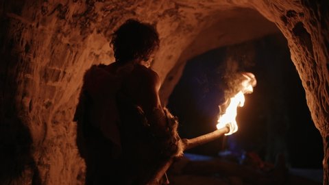 Primeval Caveman Wearing Animal Skin Exploring Cave At Night, Holding Torch with Fire Looking at Drawings on the Walls at Night. Neanderthal Searching Safe Place to Spend the Night.Back View Following