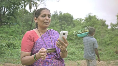 A happy female farmer/ Businesswoman making a payment holding a mobile phone and credit/debit card in her hands and a man carrying the harvested fresh fruits and vegetables