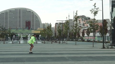 A beautiful girl from Russia is riding a skateboard along the street. Modern girl in motion. She is wearing a bright jacket with a light green hood.