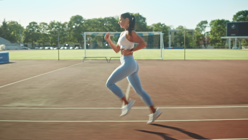 Beautiful Fitness Woman in Light Blue Athletic Top and Leggings Jogging in the Stadium. She is Running Fast on a Warm Summer Afternoon. Athlete Doing Her Routine Sports Practice. Tracking Shot.