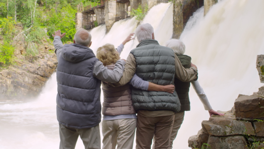 Handheld shot with rear view of senior people standing together in front of dam spillway and discussing view