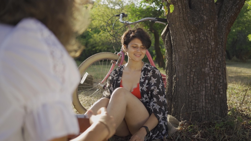 Attractive young woman with short hair sitting under the tree while her friend taking photo in the garden. The bicycle standing behind. Rural life. Retro style