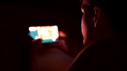 Teenage boy playing a video game on his smartphone in the darkness at night. UHD
