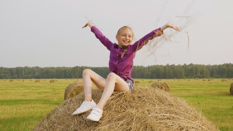Girl scattering straw on field. Adorable happy girl in checkered shirt and denim shorts sitting on haystack and smiling at camera. Harvest concept