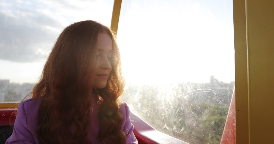 Young woman with curly long red hair is sitting in funicular or ferris wheel cabin in time of sunset. Natural freckles on smiling female face. Sun light flaring, lens glaring.