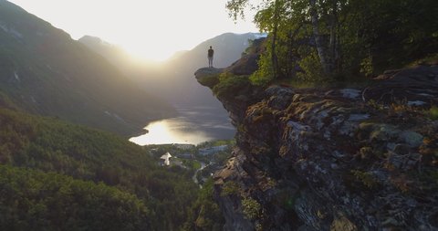Aerial: Full length of tourist with arms raised on rocky cliff against Geirangerfjord amidst mountains during sunset - Geiranger Fjord, Norway